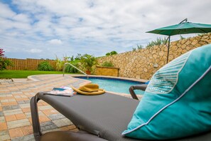Un espace aménagé au bord de la piscine, avec un fauteuil de relaxation, un chapeau et un parasol.
