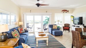 A spacious living room with a blue sofa, a coffee table, and a TV. There are two chairs, a lamp, and a ceiling fan. -  Oceanfront Marina Condo With All Luxuries (Key West)