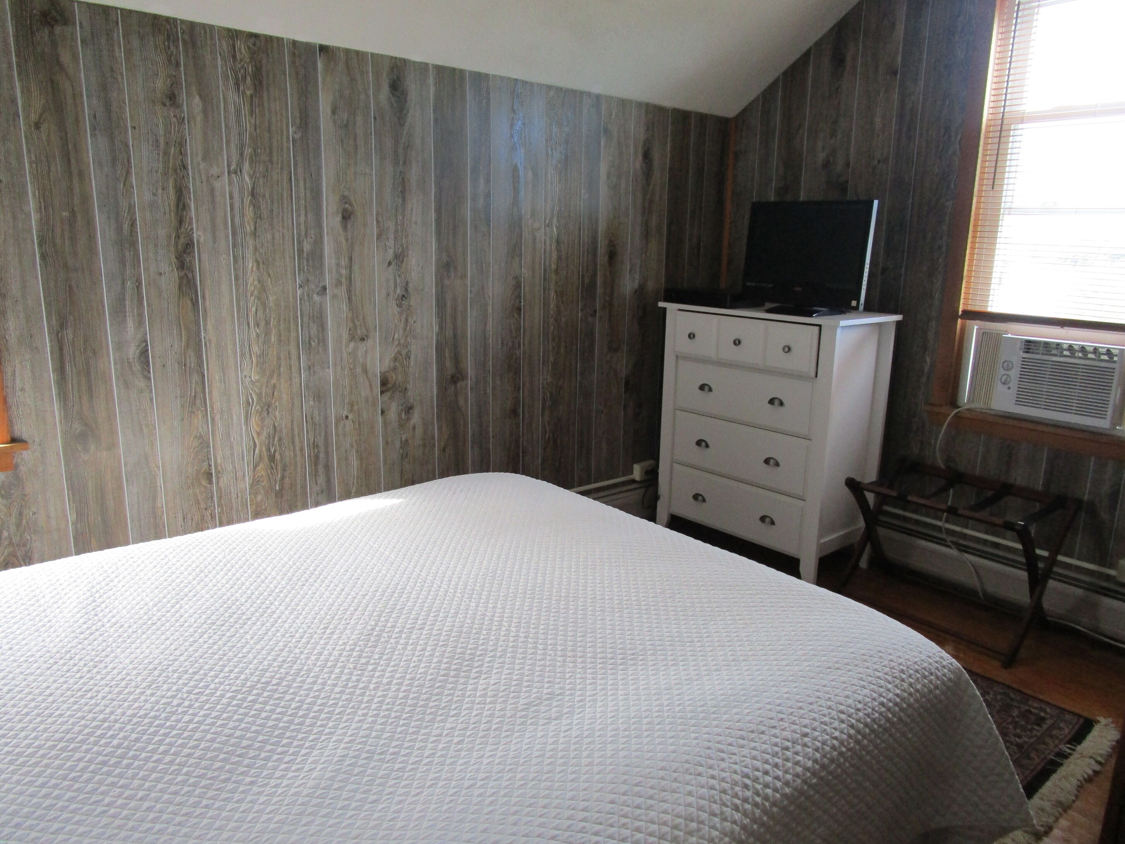 A bedroom with a bed, a dresser, a television on top of the dresser, and an air conditioning unit mounted on the wall.