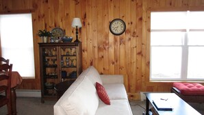 Ein Wohnzimmer mit einem weißen Sofa, einem roten Kissen, einer Holzkabine mit Gläsern, einer Uhr an der Wand und einem Fenster mit Jalousien.