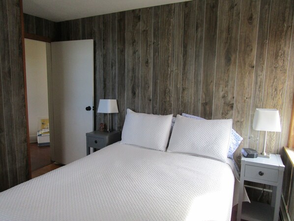 Un dormitorio con pared de madera, una cama con ropa de cama blanca, dos mesitas de noche con lámparas y una puerta a otra habitación.