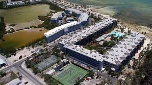 Vue aérienne d’un complexe hôtelier comprenant plusieurs bâtiments, un court de tennis et une plage.