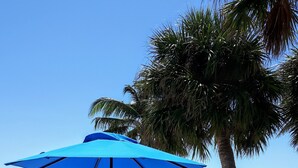 Un salon de plage avec un parasol bleu, des chaises en métal blanches et une vue sur l’océan.