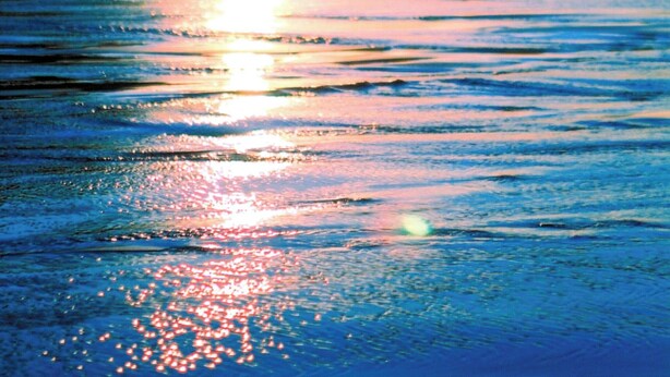 Een strandscène met zonlicht dat op het water reflecteert, kliffen op de achtergrond en een heldere lucht.