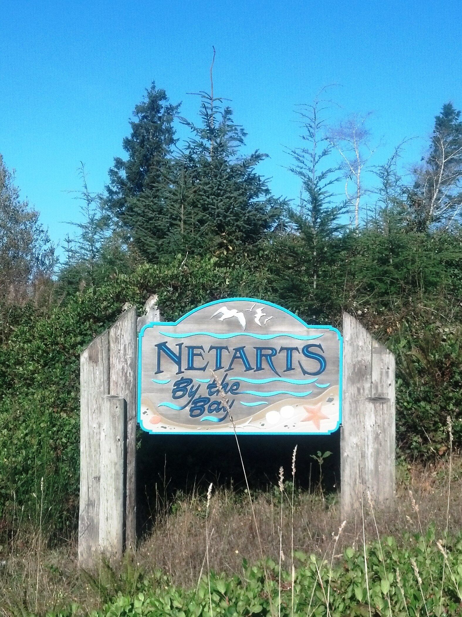 Een houten bord met de tekst 'NETARTS By the Bay', omringd door groen.