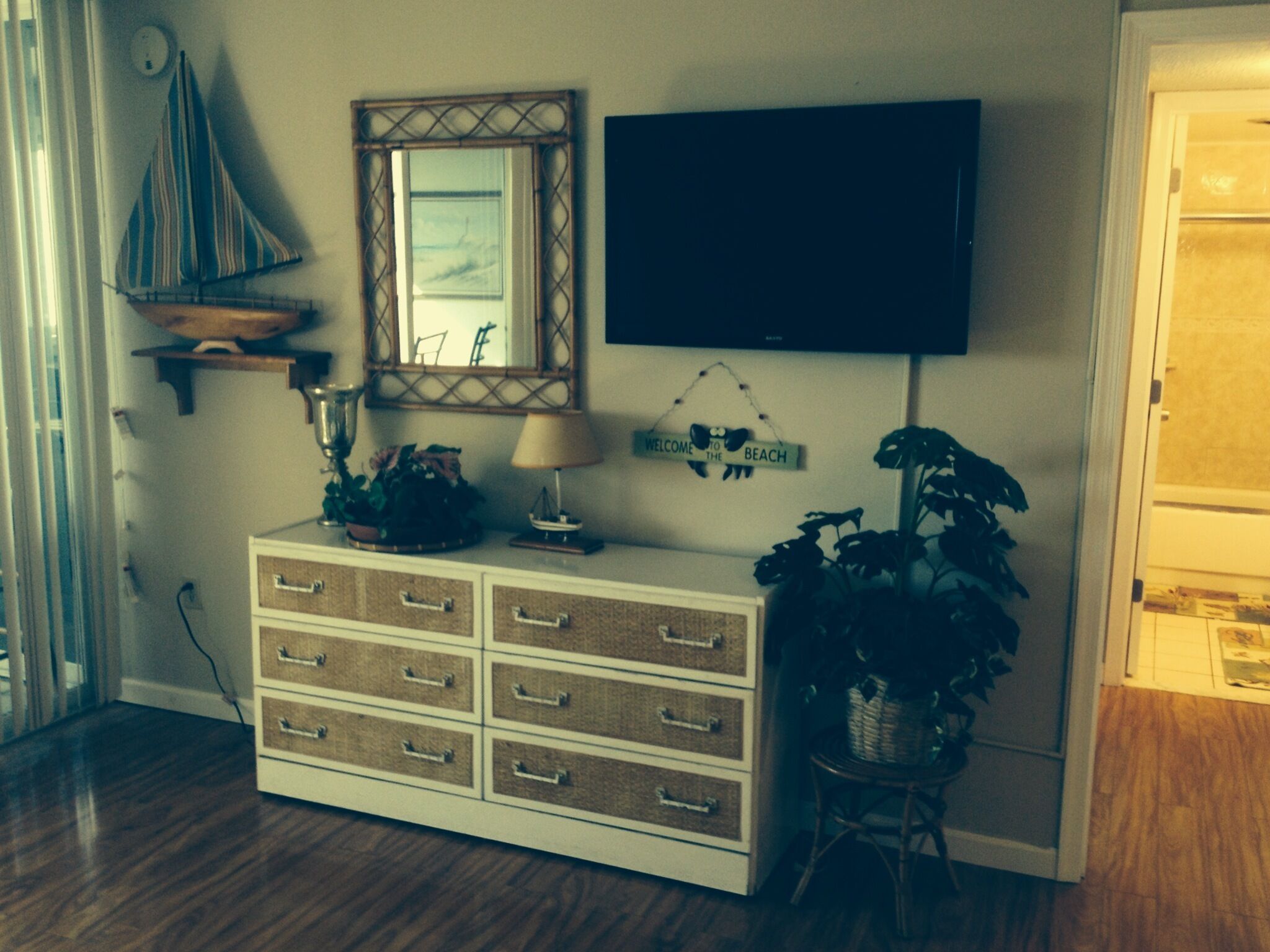 A room with a wooden dresser, a television mounted on the wall, a decorative mirror, and a welcome sign.
