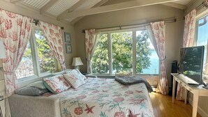 A bedroom with a wooden floor, a bed with floral bedding, a desk with a TV, and large windows with curtains.