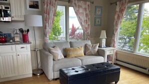 A living room with a grey sofa, two starfish pillows, a blue ottoman, a kitchen area with white cabinets, and a window with curtains.