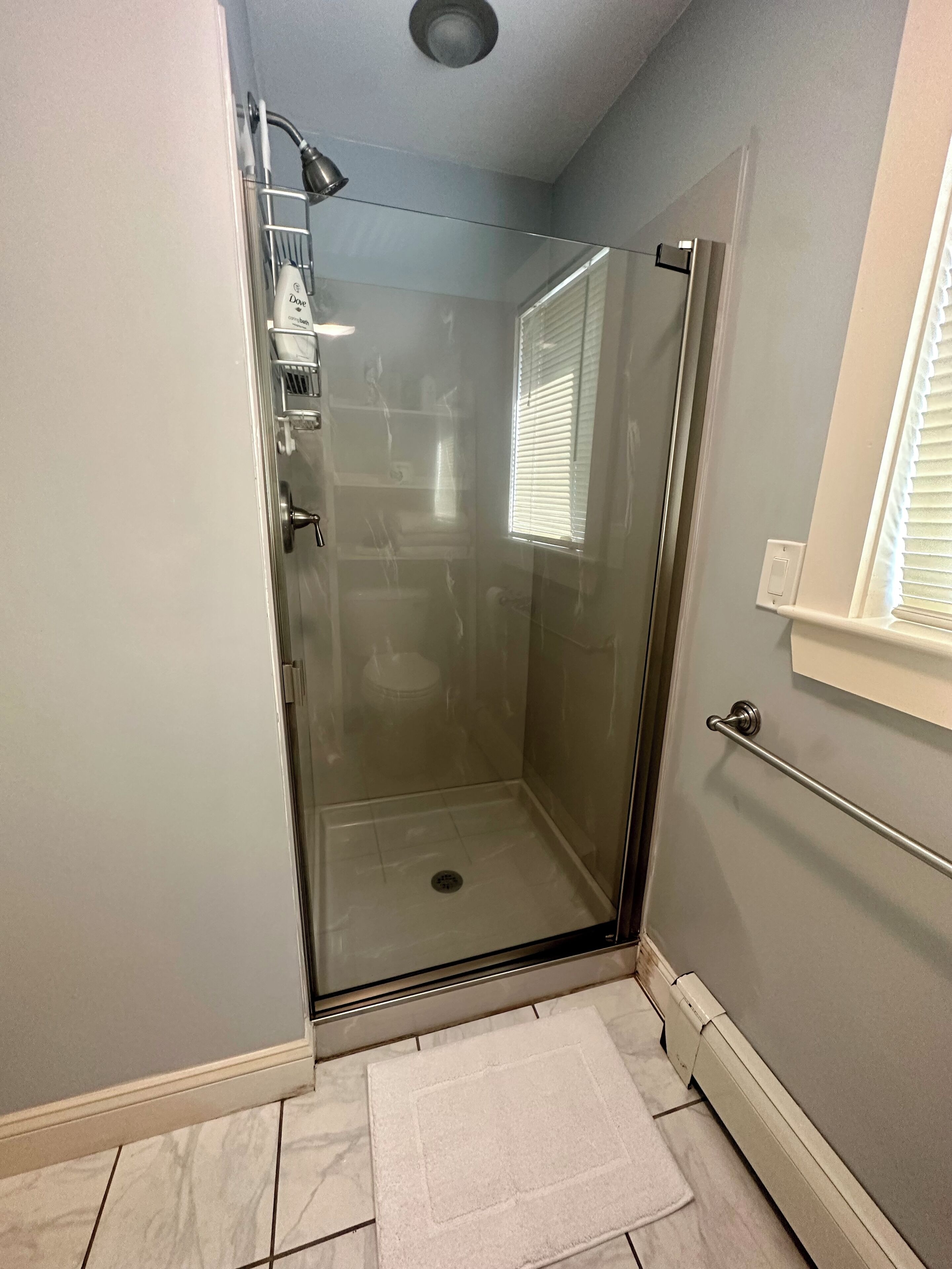Une cabine de douche fermée par des parois en verre, avec un tapis de sol blanc, un porte-serviettes et une fenêtre équipée de stores.