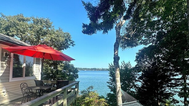 A wooden deck with outdoor furniture and a red umbrella, overlooking a lake.