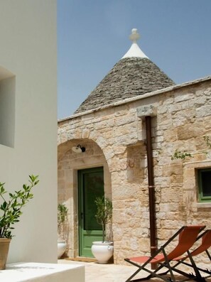 A stone building with a conical roof, a green door, and a small window. There are two potted plants and two chairs outside. - Trullo Monte Olimpo 5 (Alberobello)