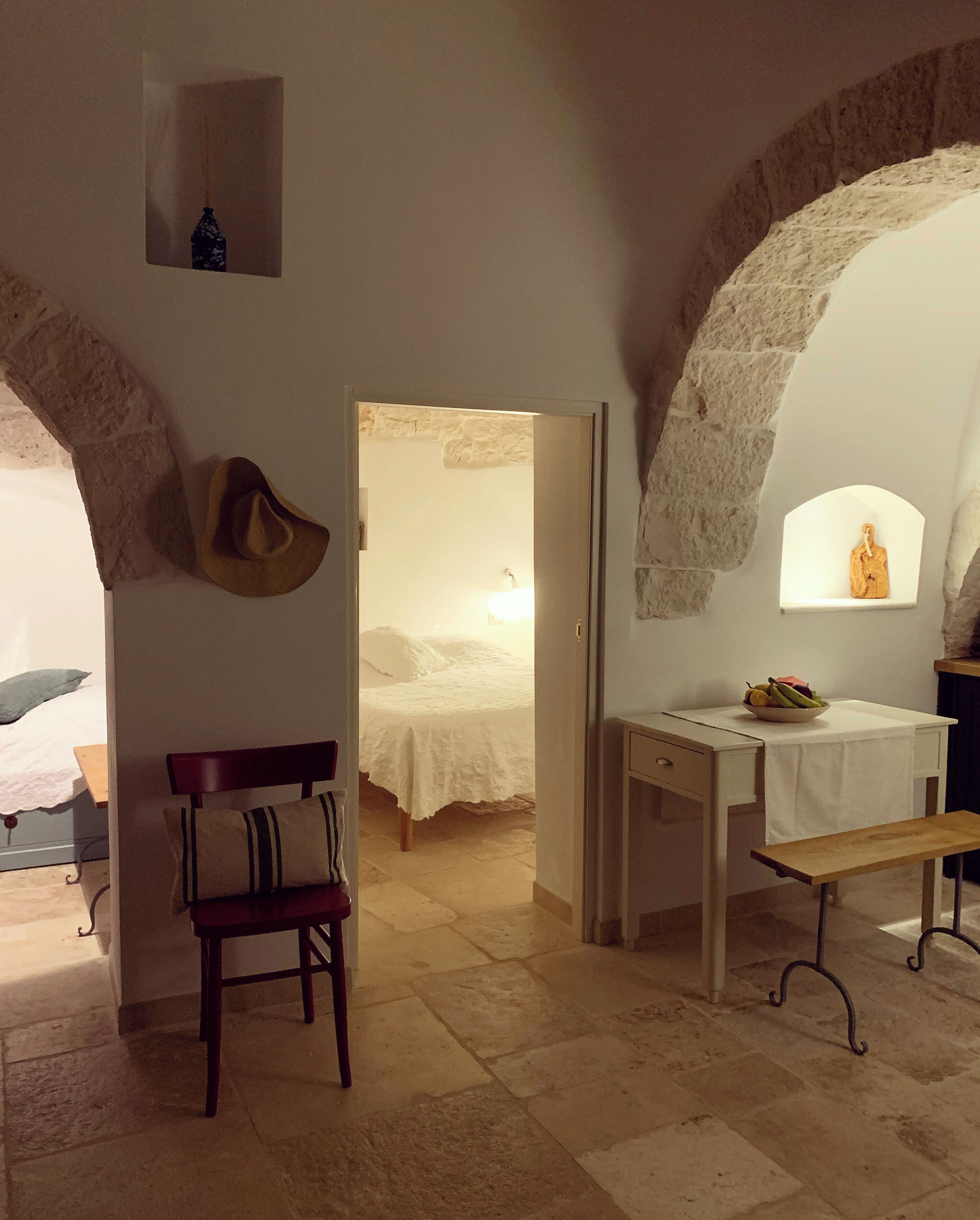 A room with a bed, a desk, a chair, and a small table with a bowl of fruit. There are stone arches and a statue on the wall.