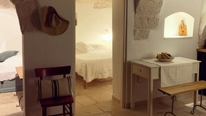 Una habitación con cama, escritorio, silla y una pequeña mesa con un frutero. Se observan arcos de piedra y una estatua en la pared.