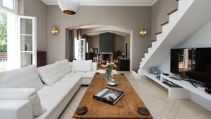 Un salon moderne avec un canapé blanc, une table basse en bois sur laquelle repose un magazine, un téléviseur posé sur un meuble blanc et un escalier avec un téléviseur sur la plateforme.