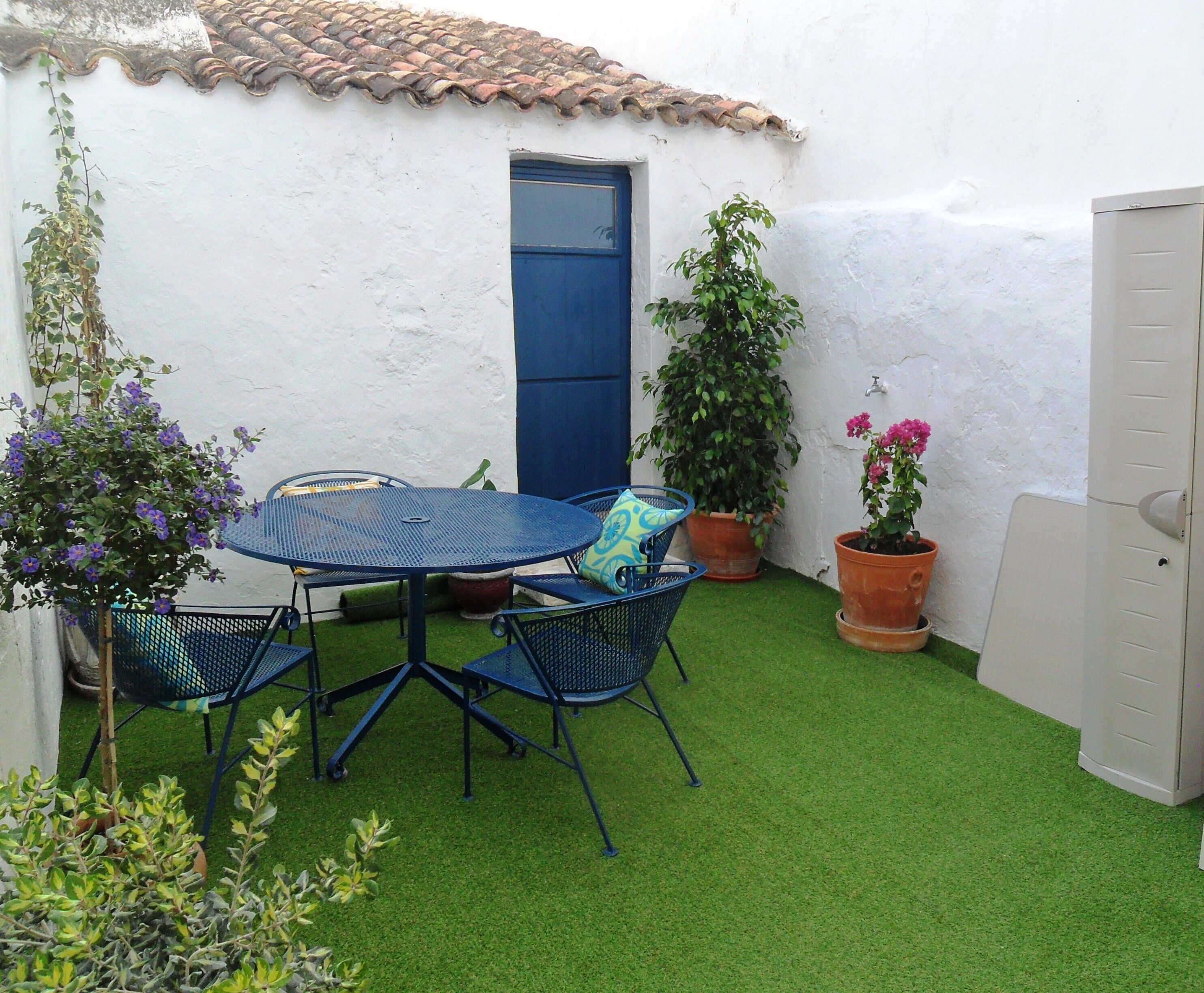A small patio with a round metal table and blue chairs, surrounded by potted plants and a white wall with a blue door.
