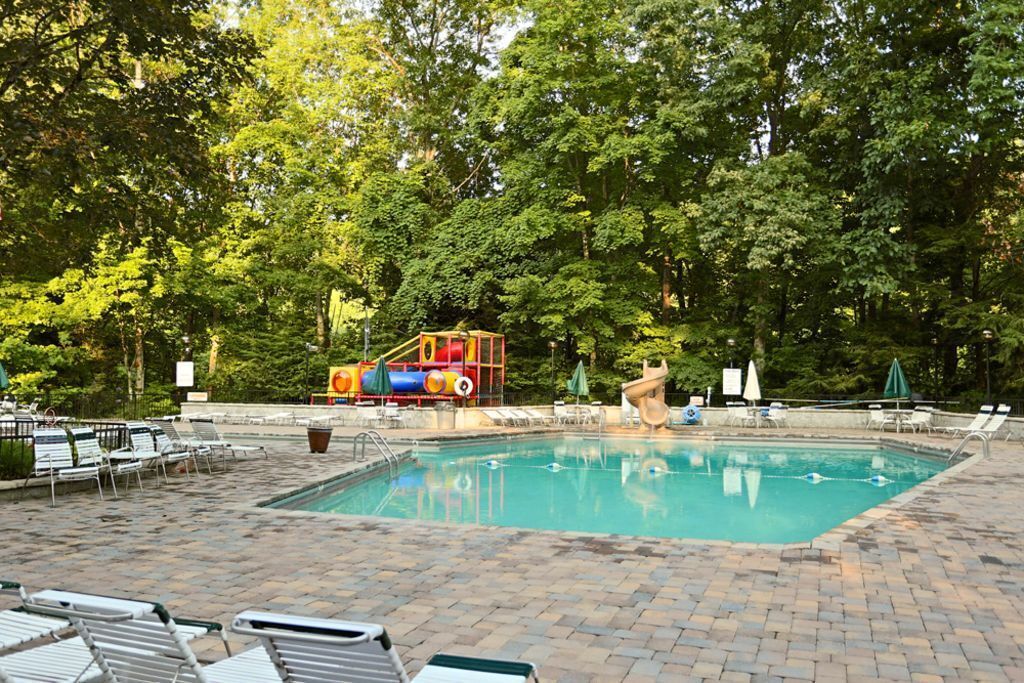 A swimming pool with a slide, surrounded by lounge chairs and umbrellas, set in a wooded area.