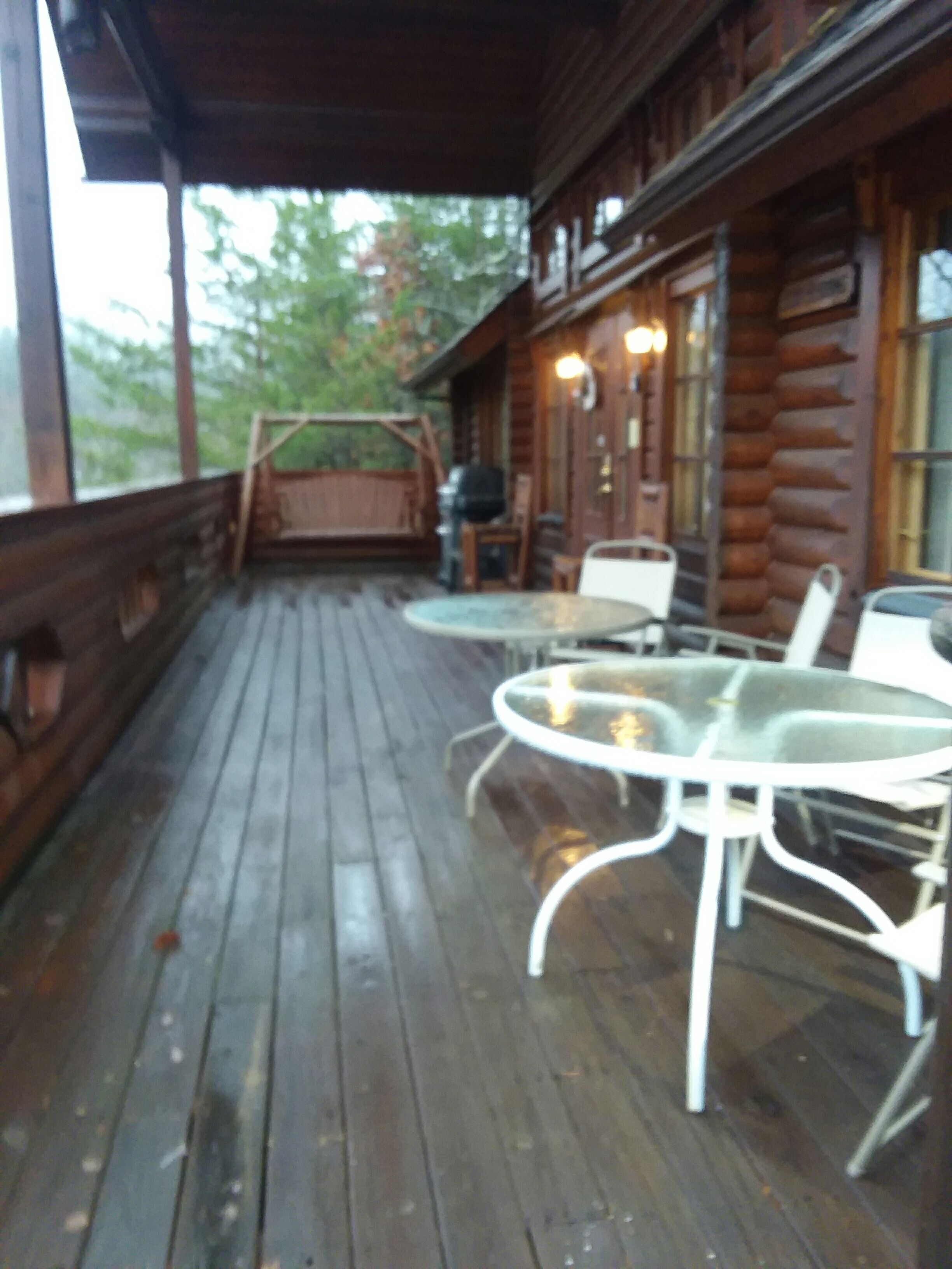 Una terraza de madera con una mesa y sillas de vidrio, un columpio y vistas al bosque.