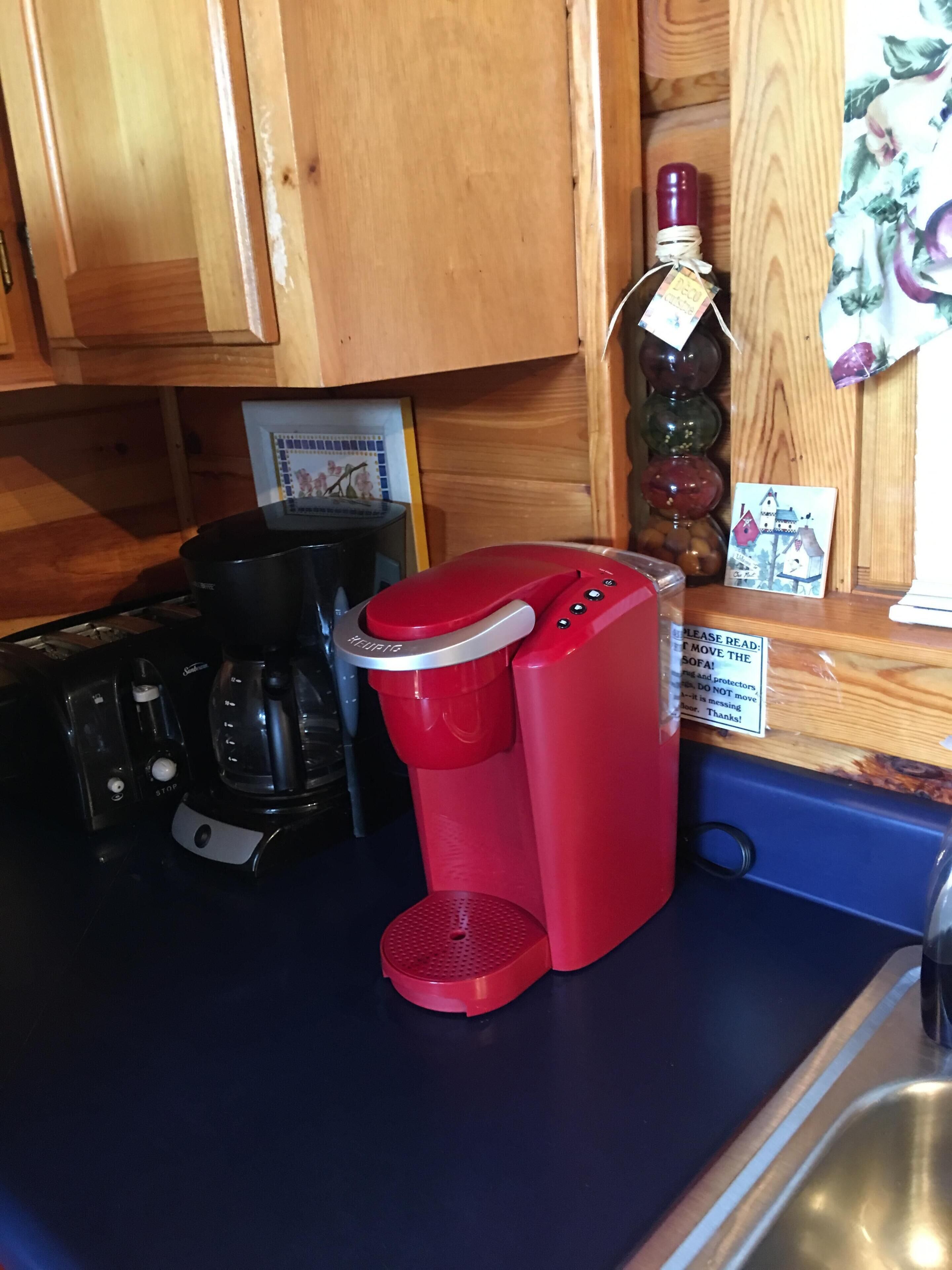Una cocina con una cafetera roja, una cafetera negra y un tostador. Hay un armario de madera sobre la encimera y un cartel en la encimera.