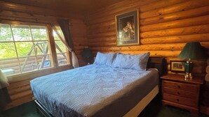Un dormitorio de cabaña de madera con cama, mesita de noche, lámpara, un cuadro enmarcado y ventana con vista a árboles.