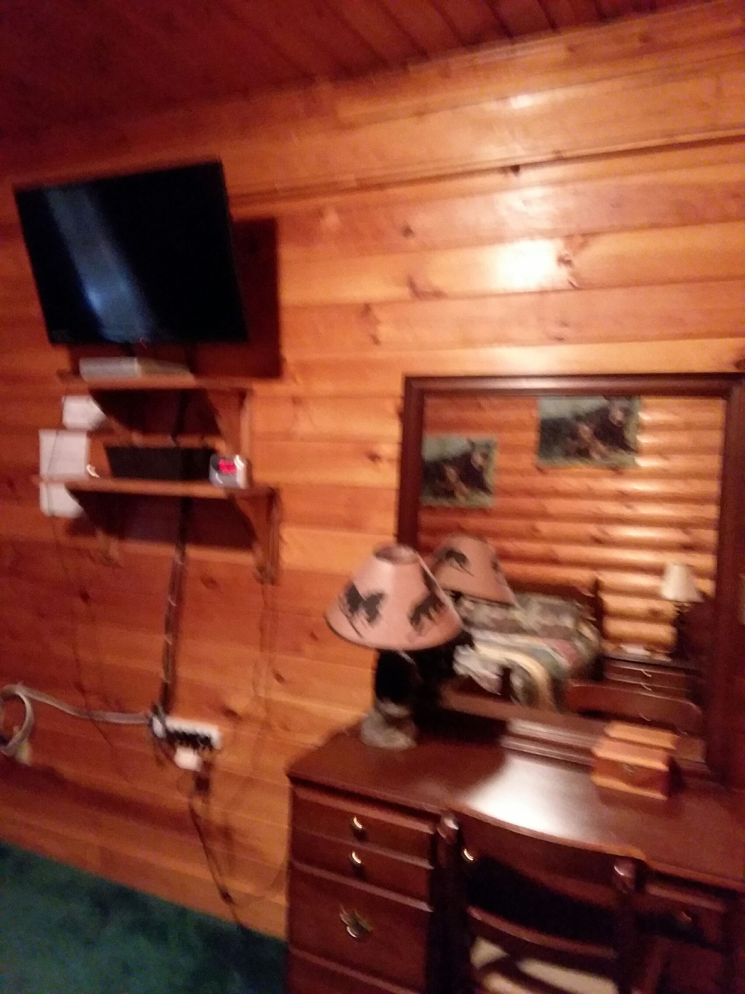 A wooden cabin interior with a television mounted on a shelf, a dresser with drawers, and a mirror with a picture frame.