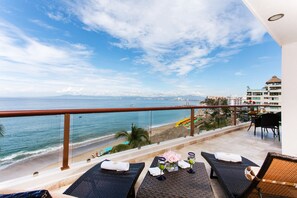 A balcony with two lounge chairs, a table with a vase of flowers, and a view of the beach and ocean. - Best location right on los muertos beach, Sky Suite E (Amapas)