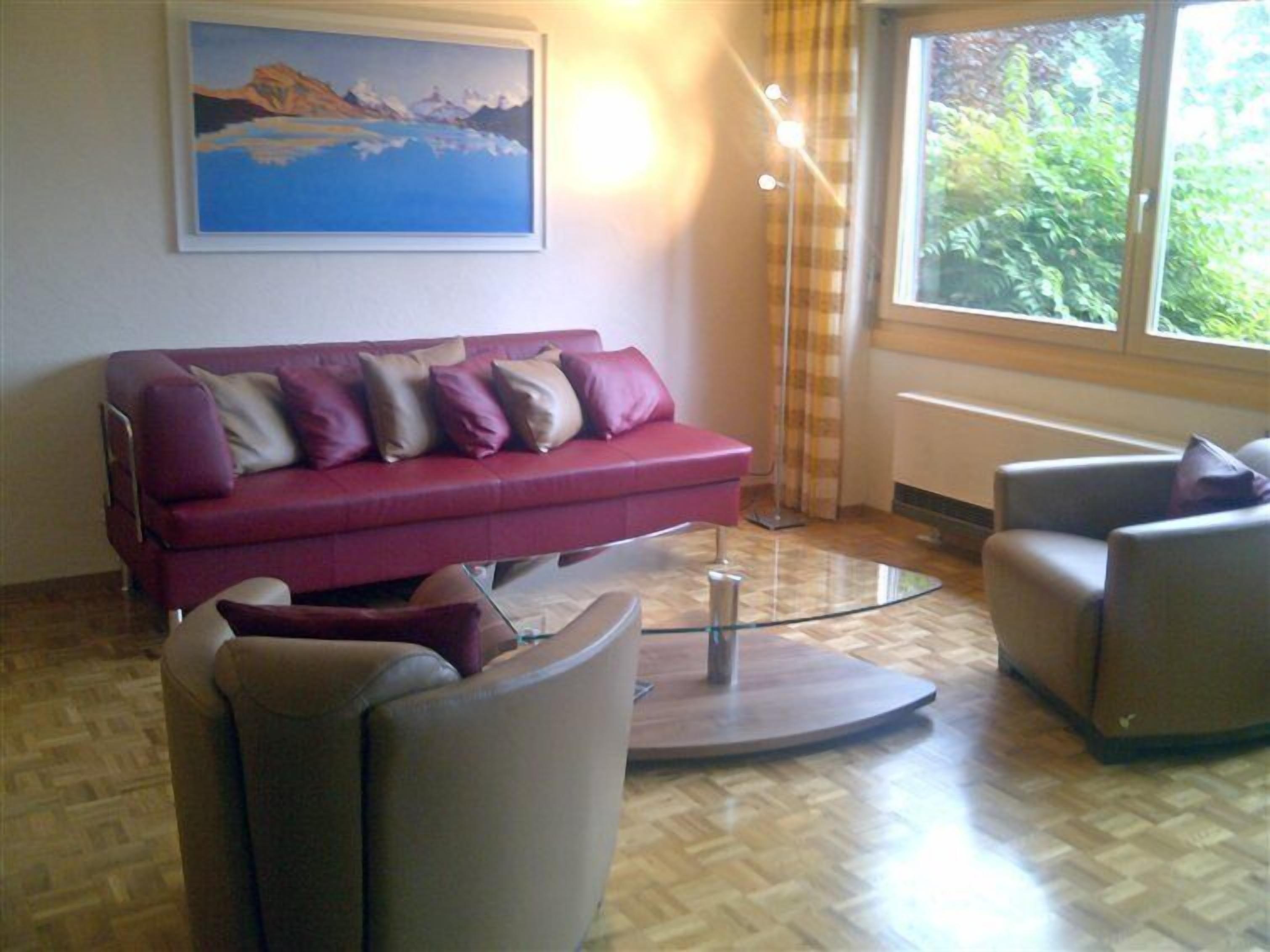A living room with a red sofa, a glass coffee table, and a grey armchair. A painting is on the wall, and there is a window with curtains.