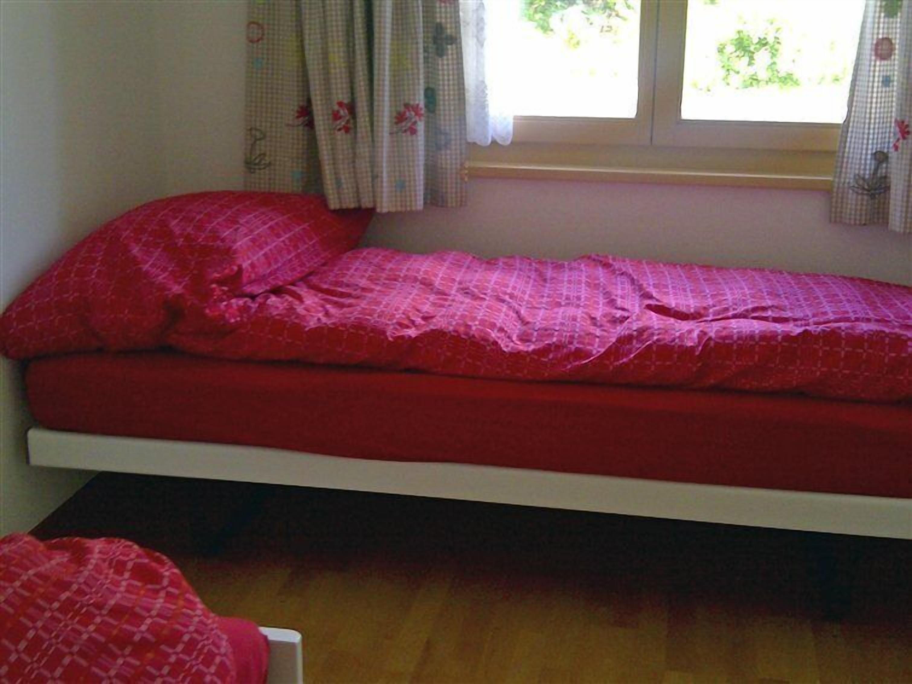 A small, single-bed room with a wooden floor, a window with floral curtains, and a red bedspread.