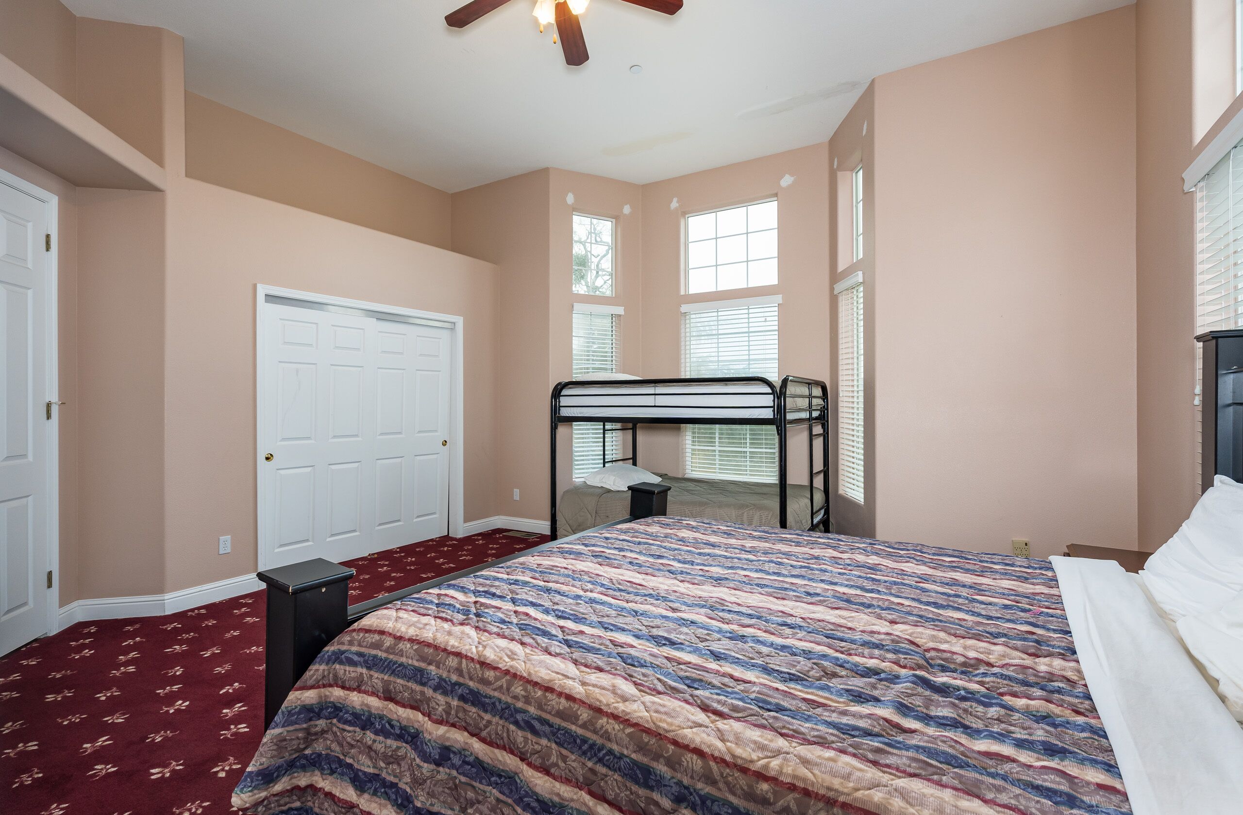 A bedroom with a bunk bed, a red carpet, a white door, and windows with blinds.