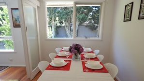 Un coin repas avec une table blanche dressée pour six personnes, des sets de table rouges, des chaises blanches et un vase rempli de fleurs violettes.
