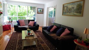 Un salon avec deux canapés noirs, une table basse et un tapis à motifs. De grandes fenêtres offrent une vue sur la verdure à l’extérieur.