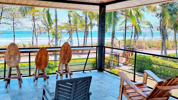 A covered outdoor area with wooden chairs and tables, overlooking a beach with palm trees and the ocean.