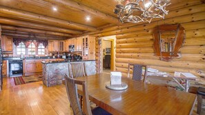 A log cabin interior with a wooden dining table, chairs, a kitchen with wooden cabinets, and a stone accent wall.