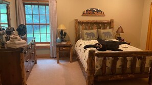 A cozy bedroom with a wooden bed, a nightstand with a lamp, a dresser, and a window with curtains.