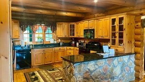 A log cabin kitchen with wooden cabinets, a stone island, and a view of the outdoors through large windows. - Lodge at Shady Branch (Hendersonville)