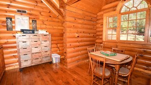 A log cabin interior with a wooden table and chairs, a window with a view of trees, and a wooden cabinet with numbered drawers.