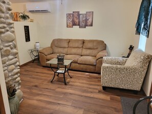 Uma sala de estar com um sofá marrom, uma mesa de centro de vidro, uma poltrona estampada, uma parede de pedra e um ar condicionado fixado na parede.