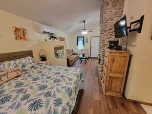 A bedroom with a bed, a nightstand, a lamp, a zebra-themed pillow, a wall-mounted TV, a stone wall, a wooden cabinet, and a small table with a chair.