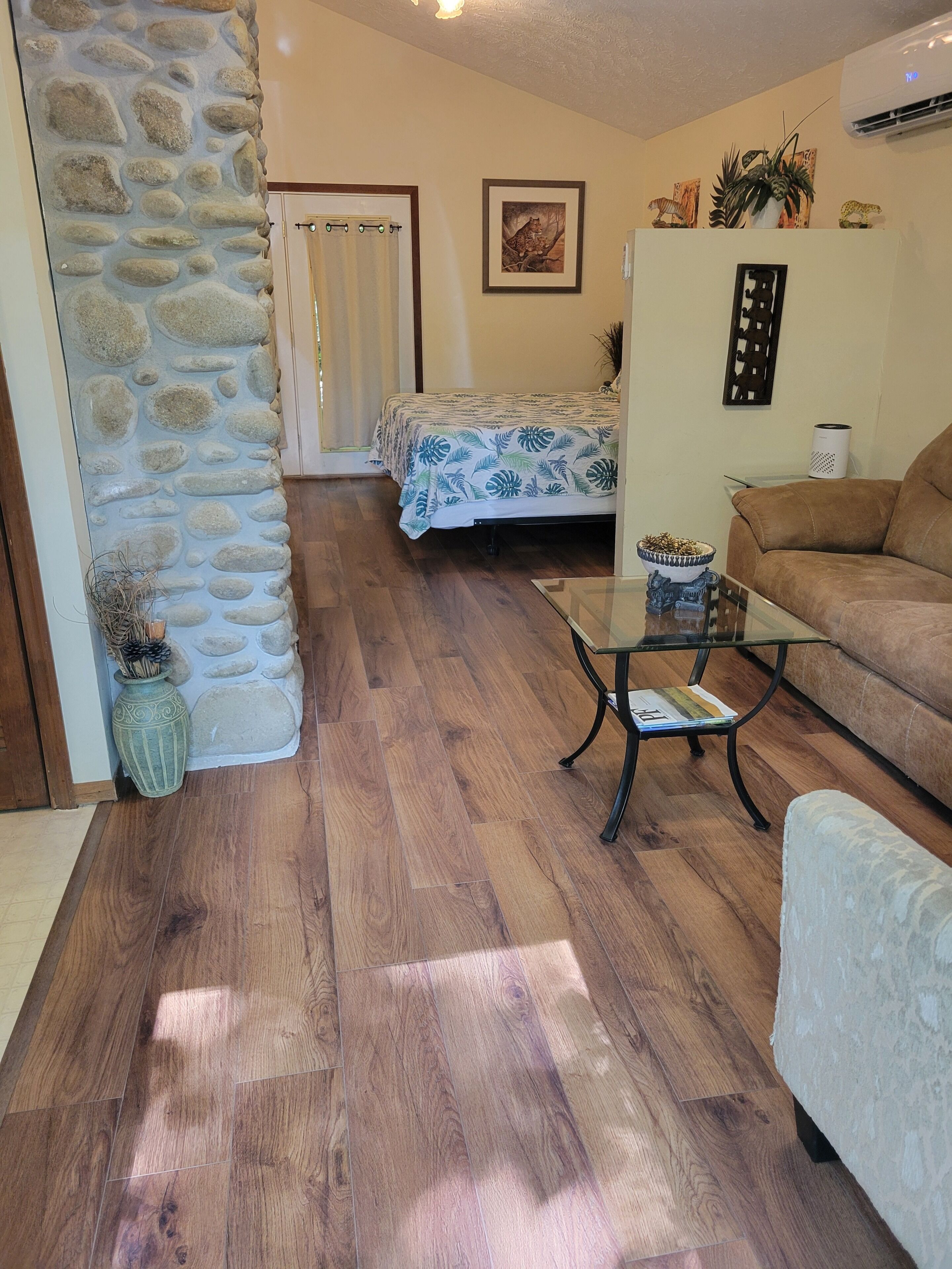 Una habitación con pared de piedra, cama con colcha estampada de flores azules, sofá marrón, mesa de centro de vidrio y una planta en maceta.