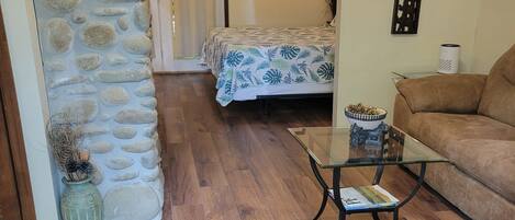 Una habitación con pared de piedra, cama con colcha estampada de flores azules, sofá marrón, mesa de centro de vidrio y una planta en maceta.