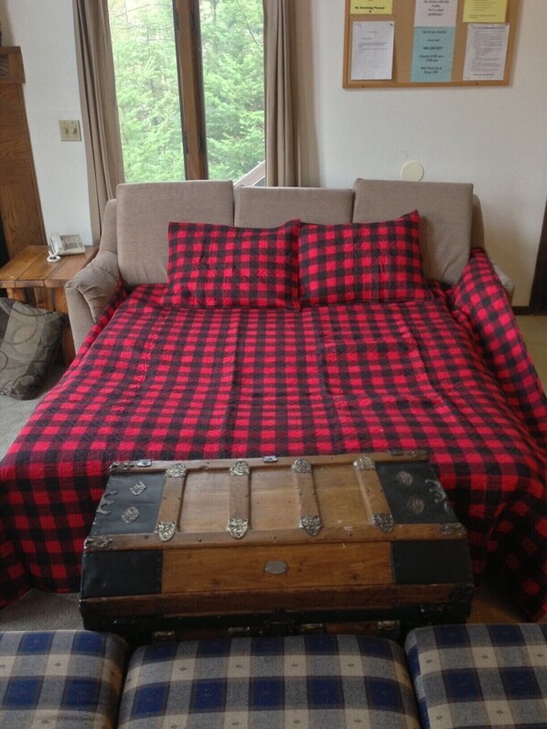 A bed with a red and black checkered bedspread, a wooden chest in front, and a window with a view of trees.
