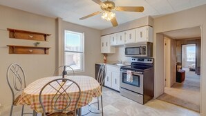 A kitchen with white cabinets, a stove, and a microwave. There is a ceiling fan with lights, a window, and a dining table with chairs.
