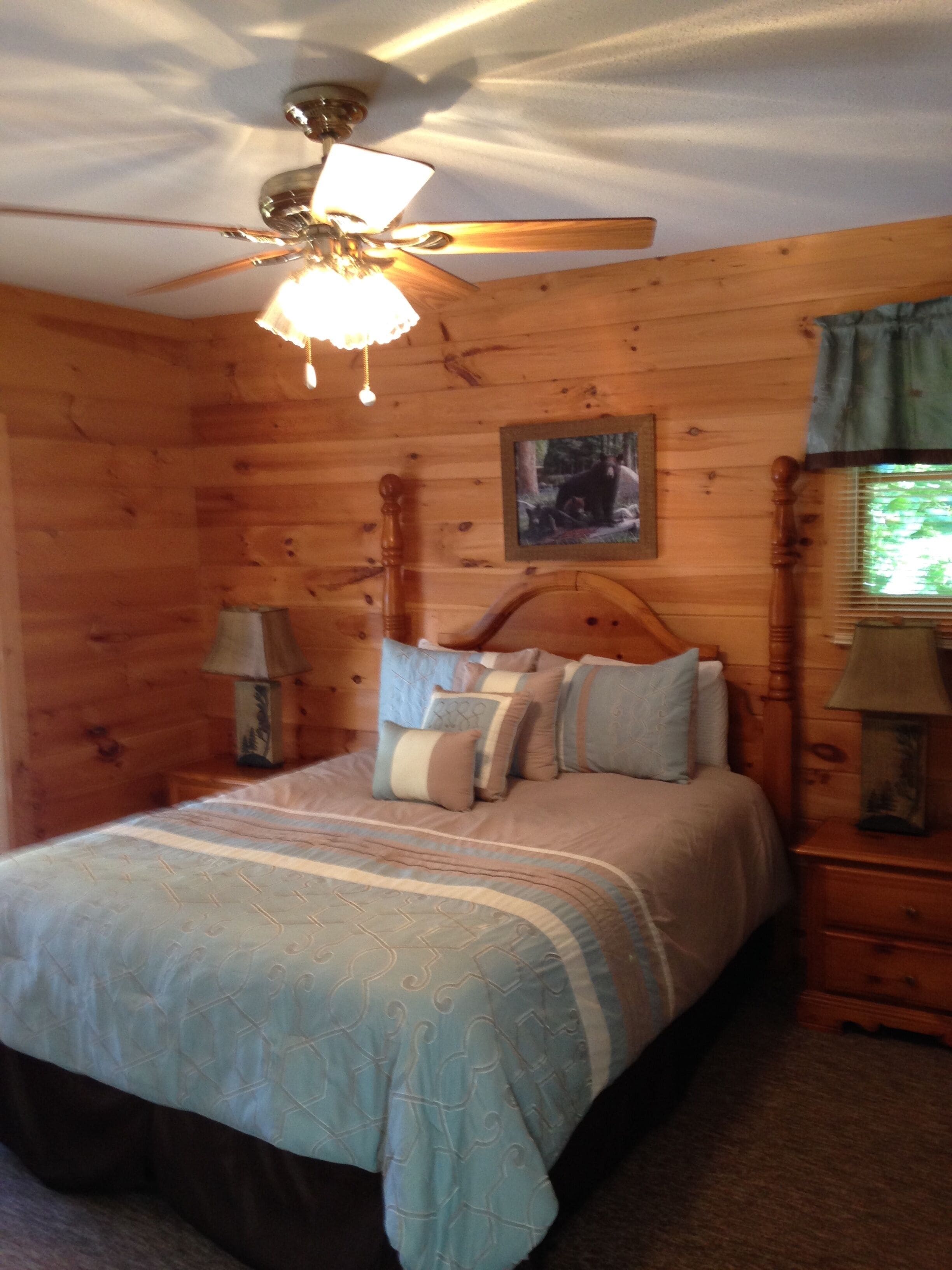 A wooden cabin bedroom with a bed, a ceiling fan, a framed picture on the wall, and a window with curtains.