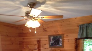 Camera da letto in una baita di legno, con un letto, una ventola a soffitto, un quadro appeso al muro e una finestra con tende.