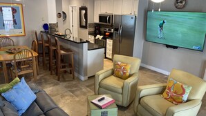 Een moderne keuken met een centraal eiland, een eethoek met houten stoelen, een bank met gekleurde kussens en een televisie waarop een golfwedstrijd wordt vertoond.
