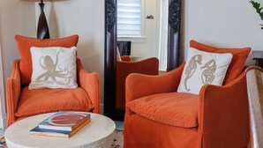Twee oranje fauteuils met decoratieve kussens, een rond bijzettafeltje met boeken en een lamp.