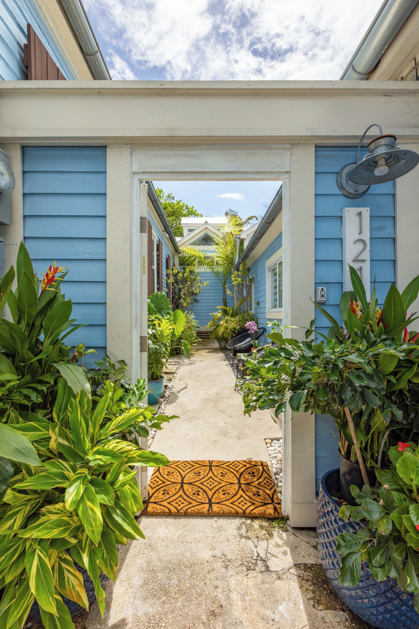 A narrow pathway leading to a house with a blue exterior, a number '12' sign, and various potted plants.