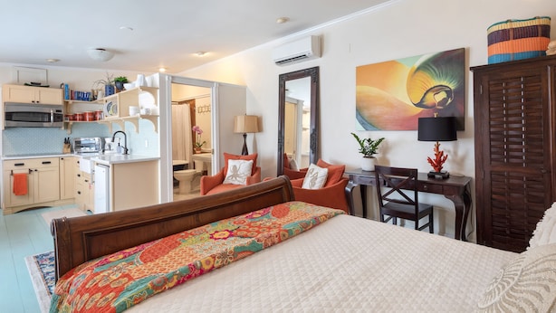 A bedroom with a large bed, a wooden headboard, a desk with a chair, a mirror, and a view of the kitchen and bathroom. - Studio Blu - Historic Key West Studio + Bikes (Key West)
