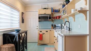 A compact kitchen with a microwave, oven, and sink. There is a small dining table with a bowl and a chair. The kitchen has light blue tiles and wooden cabinets.