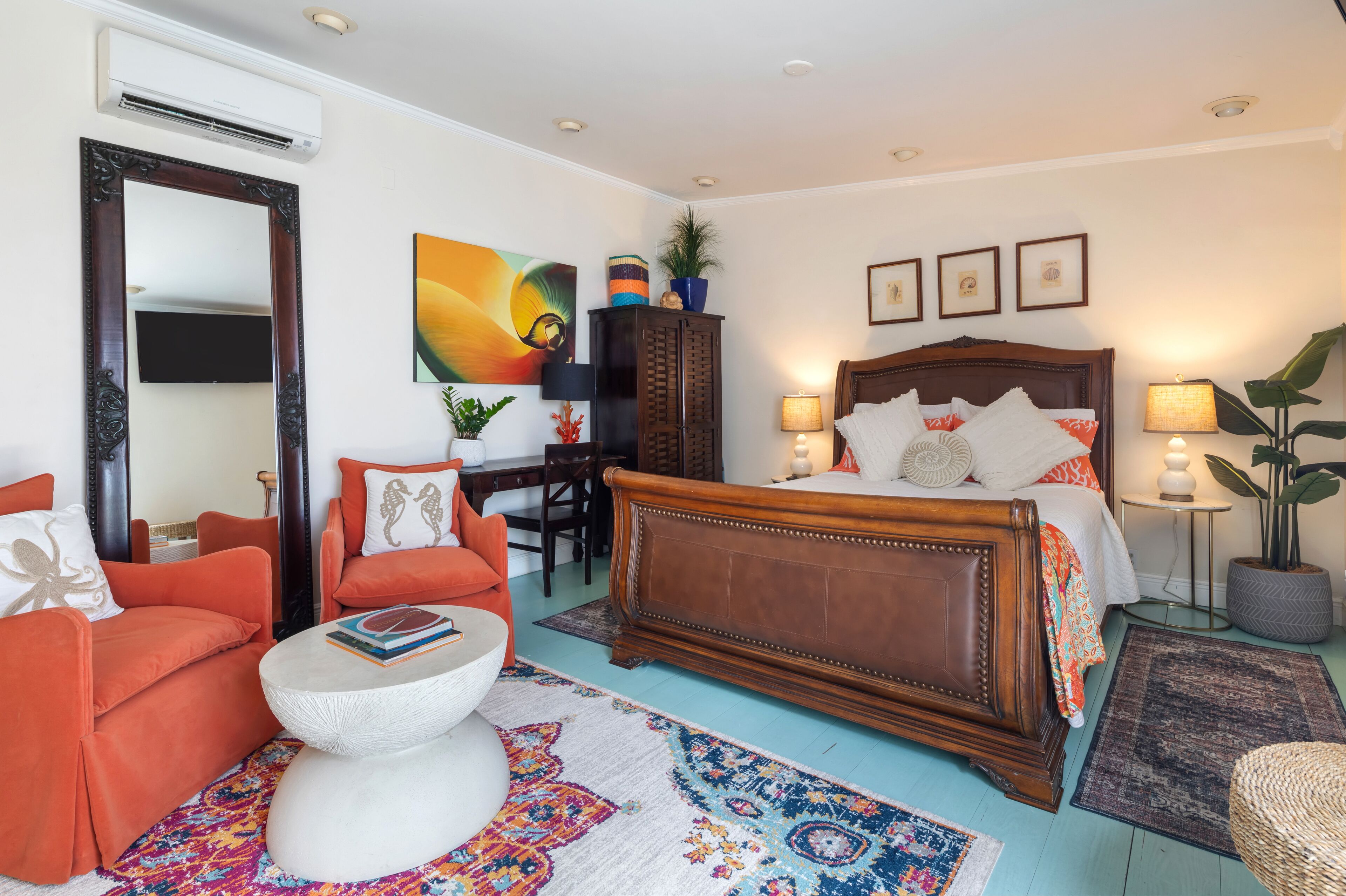 A well-lit bedroom with a wooden bed, orange armchairs, a white coffee table, and a colorful rug.