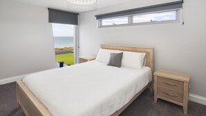Ein Schlafzimmer mit einem großen Bett, einem Nachttisch, einer Kommode, einem Fenster mit Meerblick und einer Deckenleuchte.